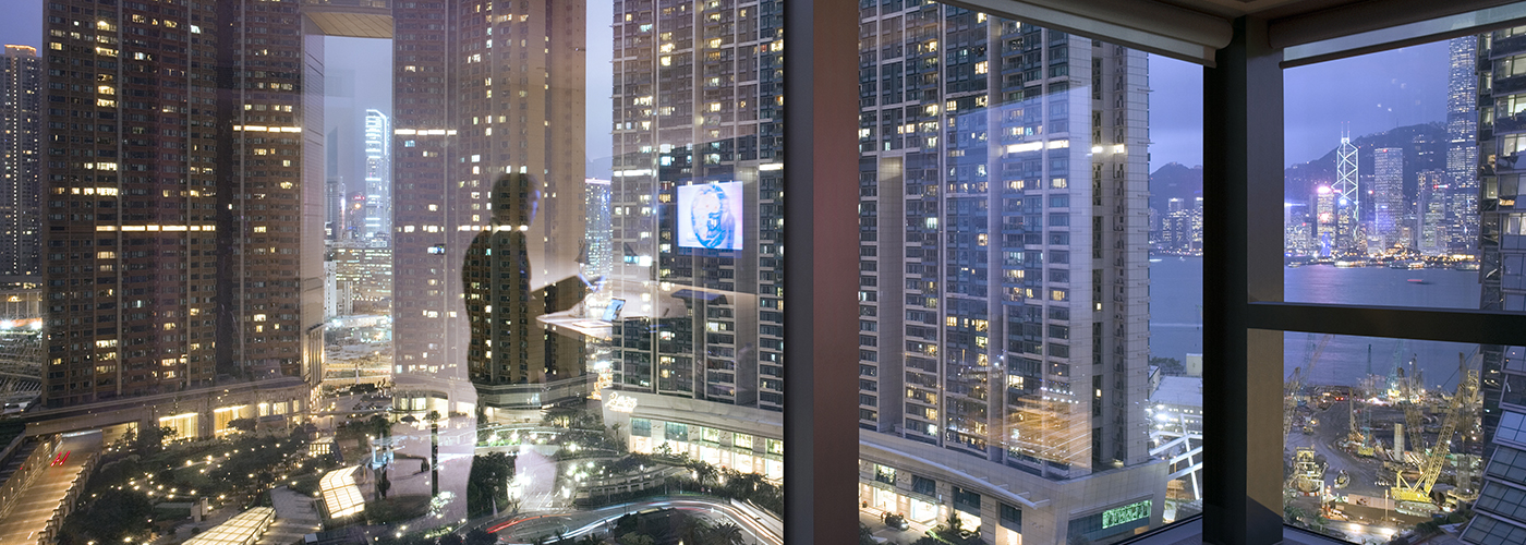 Vue panoramique depuis l’intérieur d’une pièce à travers de grandes fenêtres donnant sur un quartier d'affaires. Un homme apparaît dans le reflet de ces fenêtres.