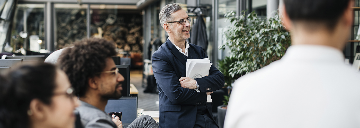 Standing man holding files, surrounded by collaborators (work environment).