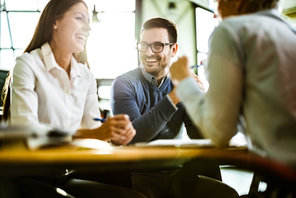 Trois personnes souriant autour d'une table (ambiance de travail détendue). 