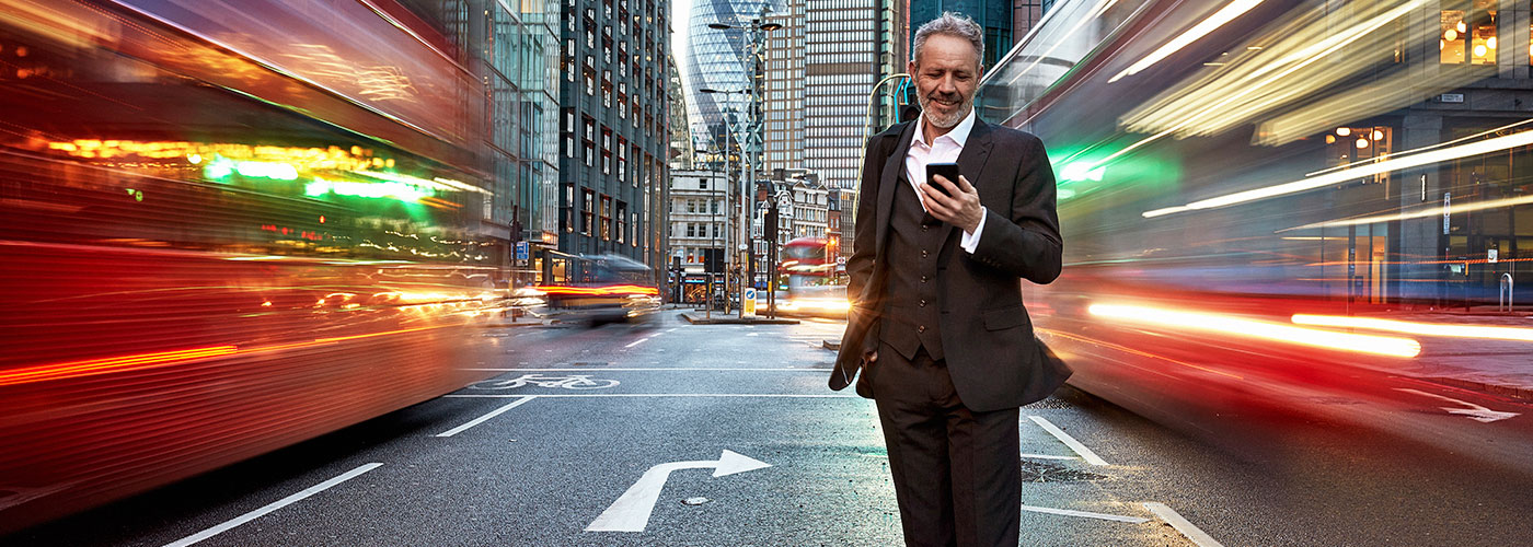 A man smiling while looking at his phone on the middle of the road with cars and buses going fast next to him.